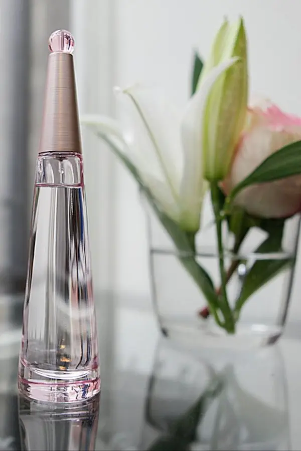 Fotografía de Producto en Madrid: Perfumes para Ecommerce y Catálogos - Sara Iglesias Primer plano de una botella de perfume de cristal con tapa rosada sobre una superficie reflectante y flores blancas de fondo, foto de fotografía de producto Madrid.